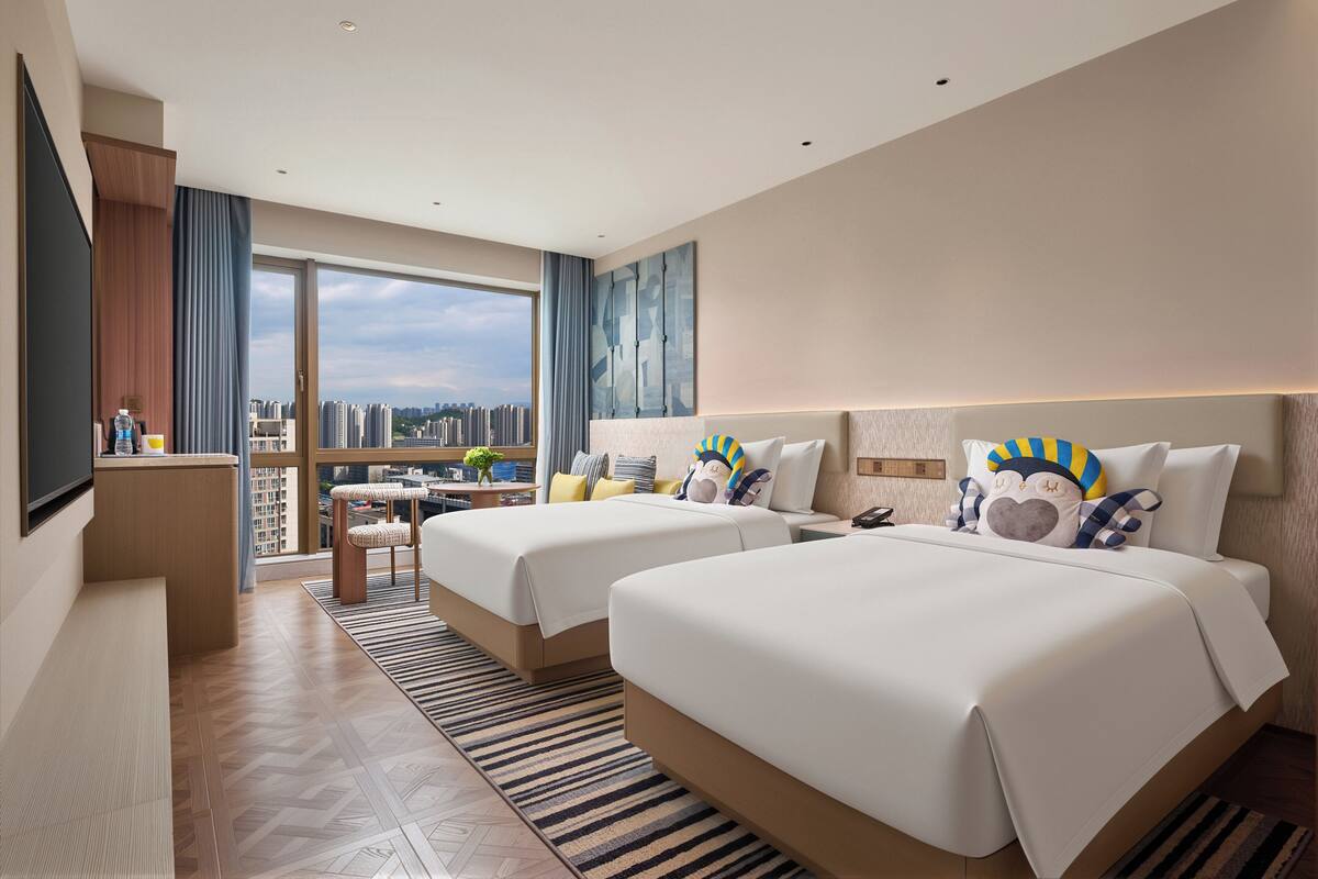 A hotel room with two beds, a desk, a TV, and a balcony with a city view.