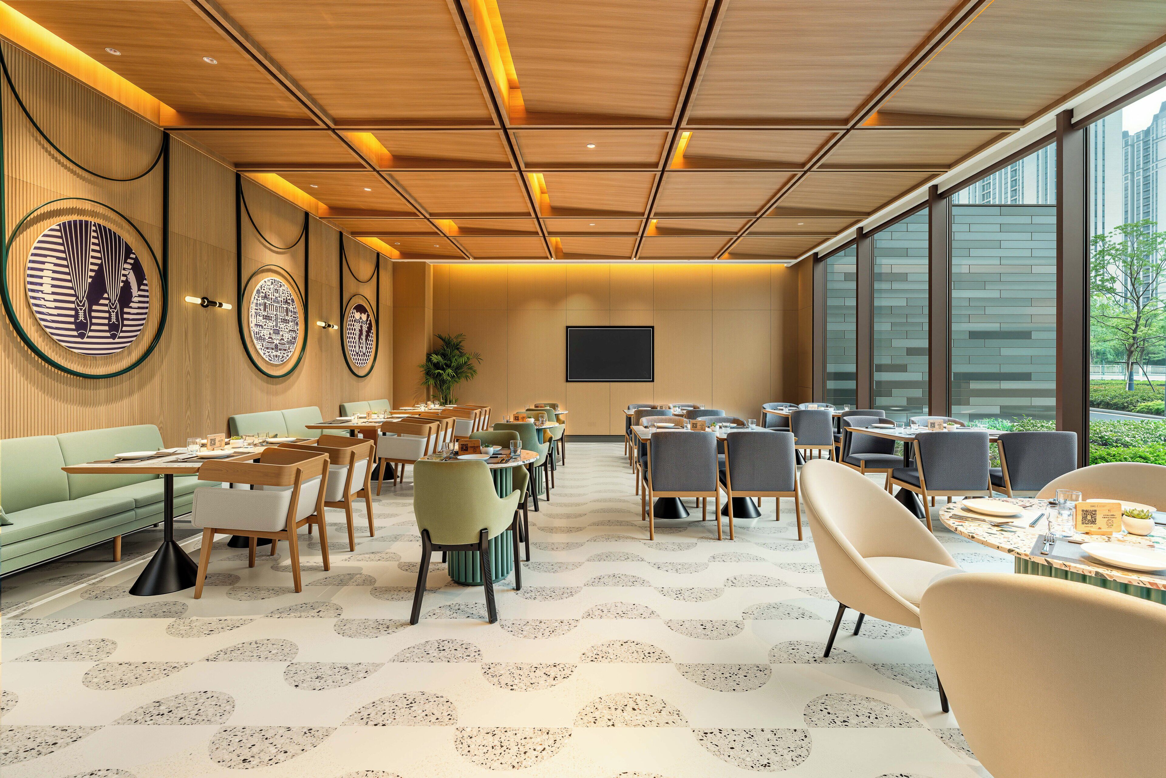 A modern hotel lobby with a wooden ceiling, circular wall art, a large TV, and a view of greenery outside.