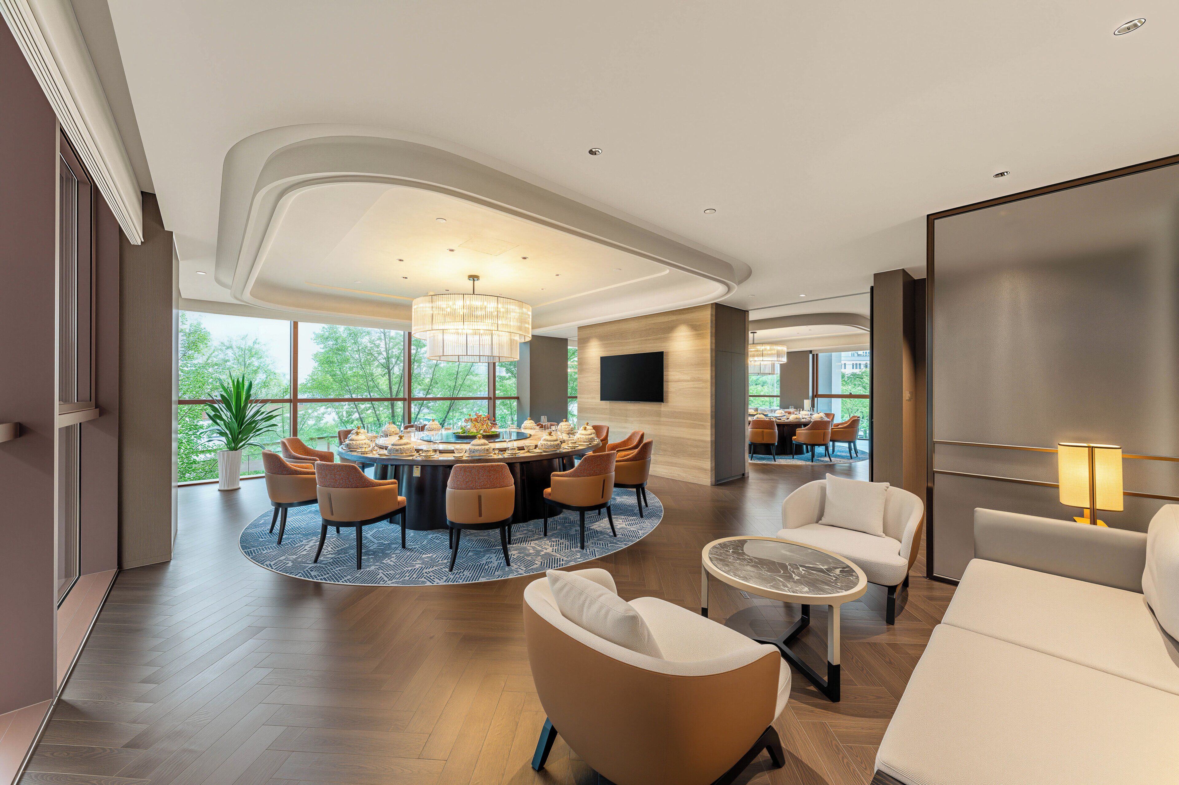 A spacious room with a round dining table, comfortable seating, and large windows offering a view of greenery.