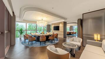 A spacious room with a round dining table, comfortable seating, and large windows offering a view of greenery.