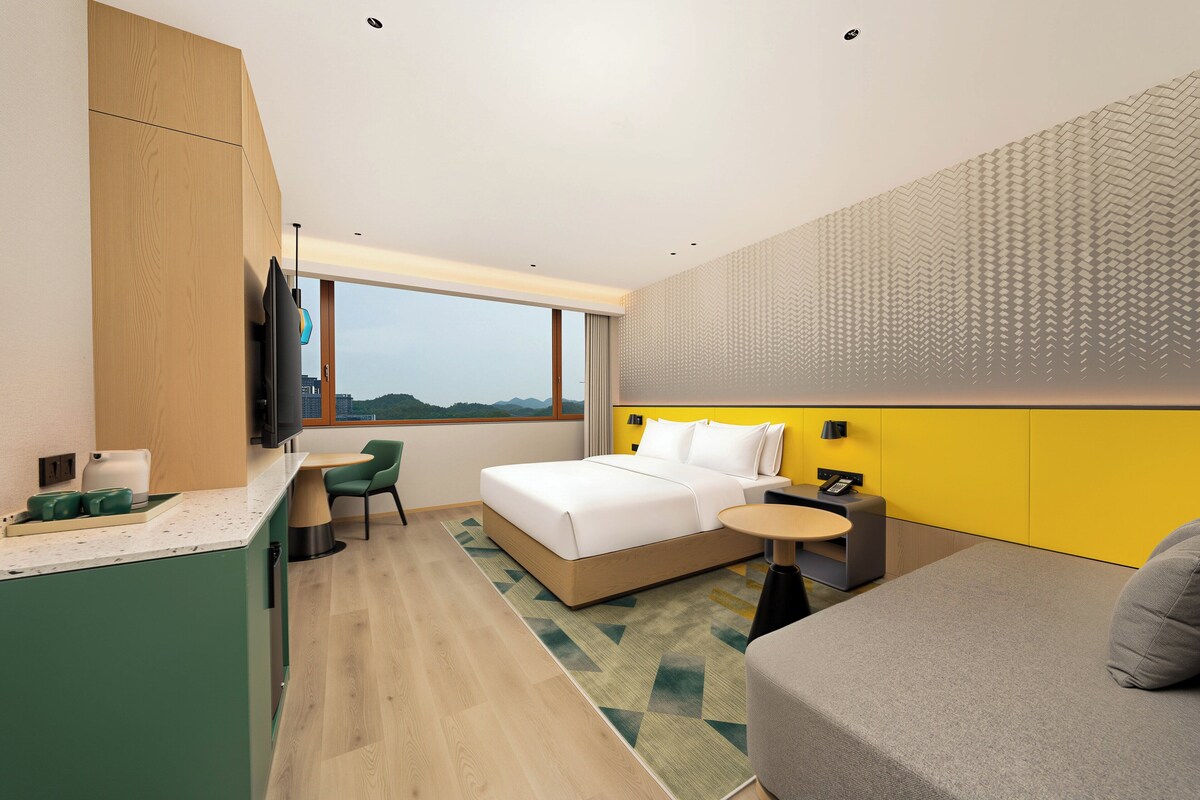 Una habitación de hotel con cama, una mesita, sofá, televisor, ventana y una zona de cocina.