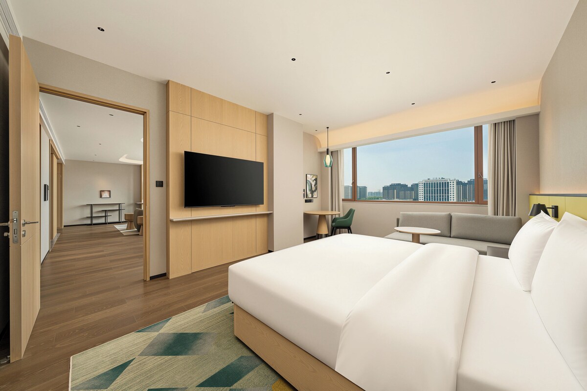 Una habitación de hotel con una cama grande, un televisor montado en la pared, un sofá, una mesita y vistas a la ciudad.