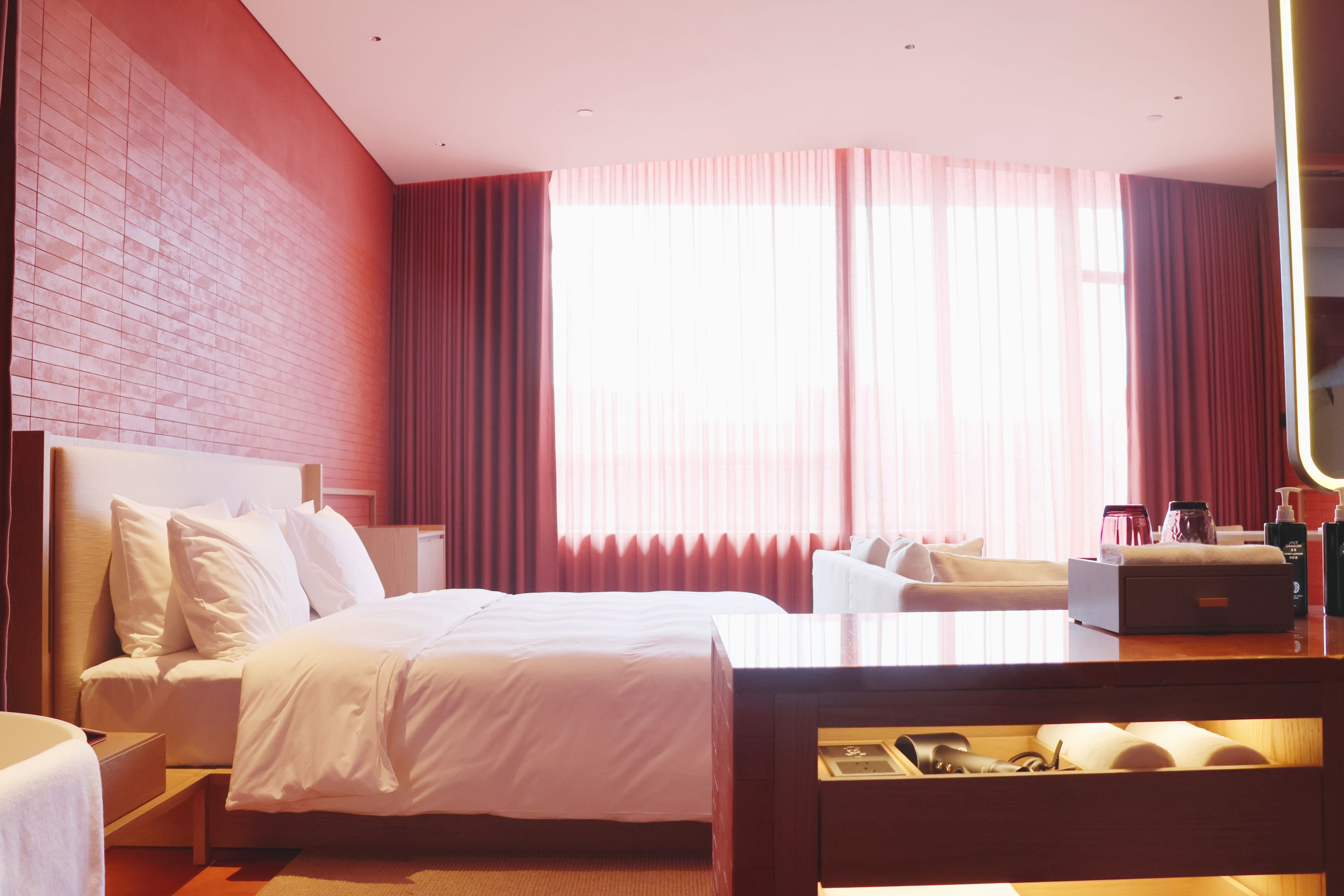 A hotel room with a large bed, a bedside table, a desk with a chair, a mirror, and a window with curtains.