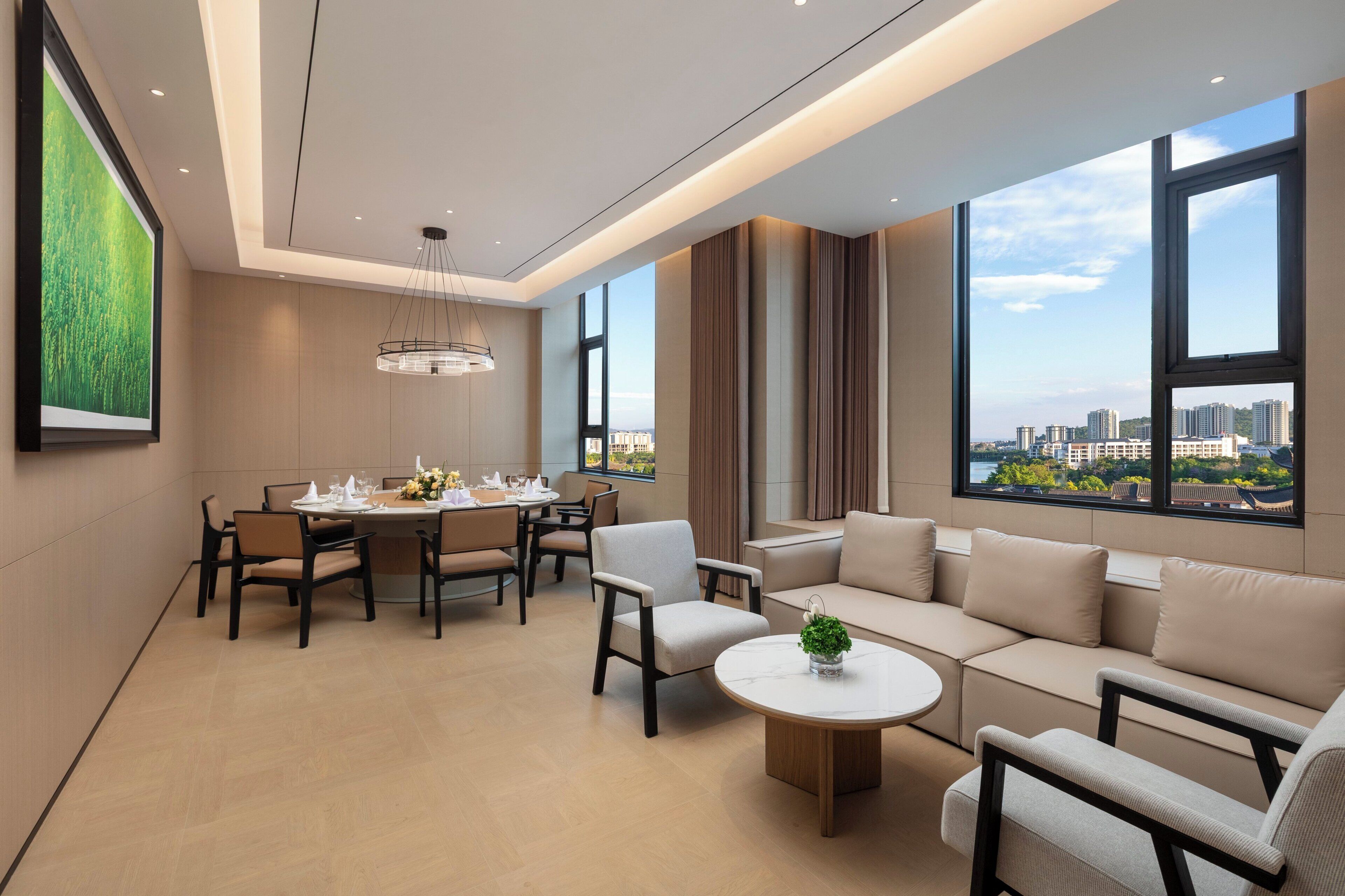 Habitación de hotel moderna con comedor, sofá, mesa de centro redonda y ventanas amplias con vistas a la ciudad.