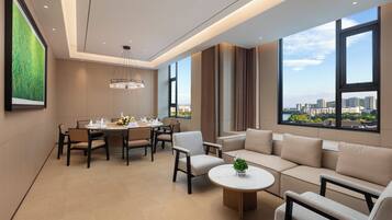 Habitación de hotel moderna con comedor, sofá, mesa de centro redonda y ventanas amplias con vistas a la ciudad.