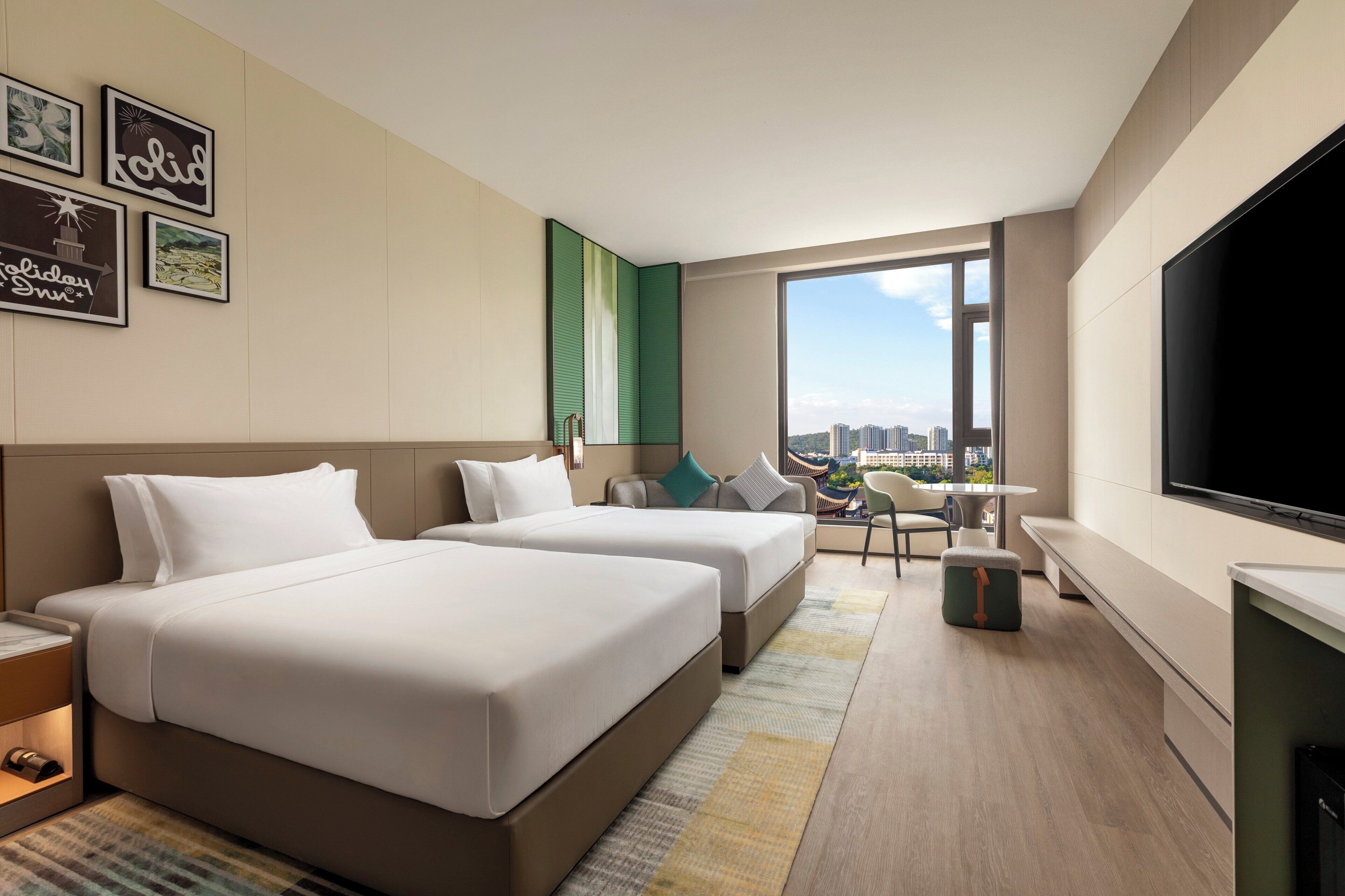 A hotel room with two beds, a TV, a balcony with a city view, and framed pictures on the wall.
