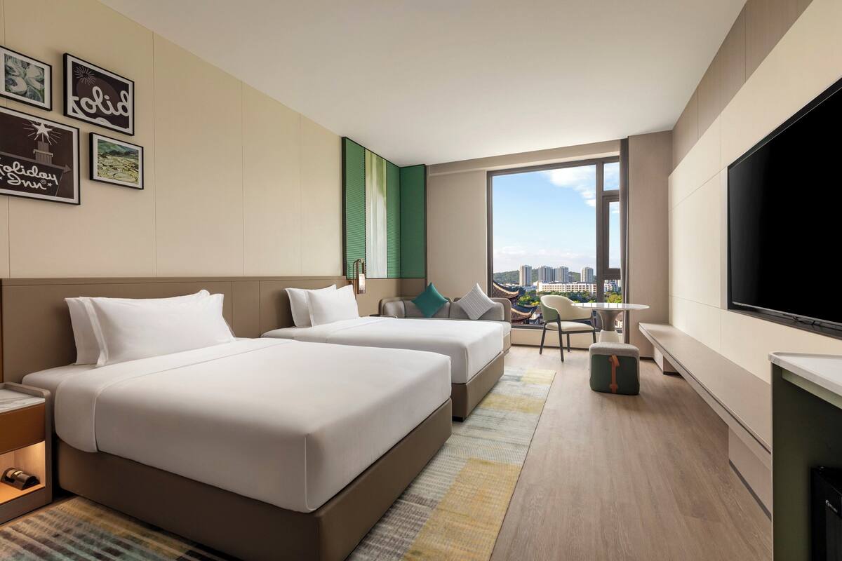 A hotel room with two beds, a TV, a balcony with a city view, and framed pictures on the wall.