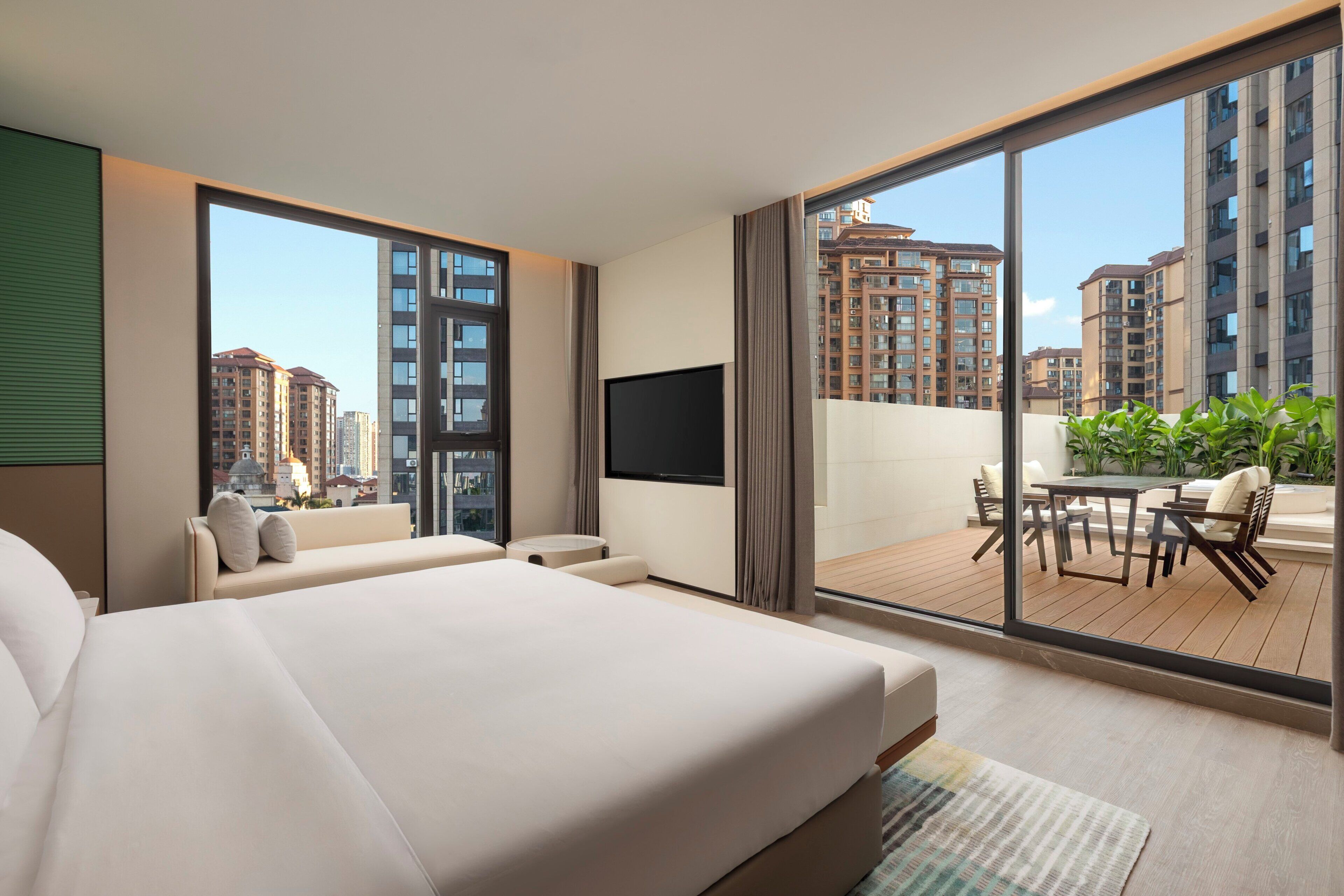 A modern hotel room with a large bed, a flat-screen TV, a balcony with a table and chairs, and a view of high-rise buildings.