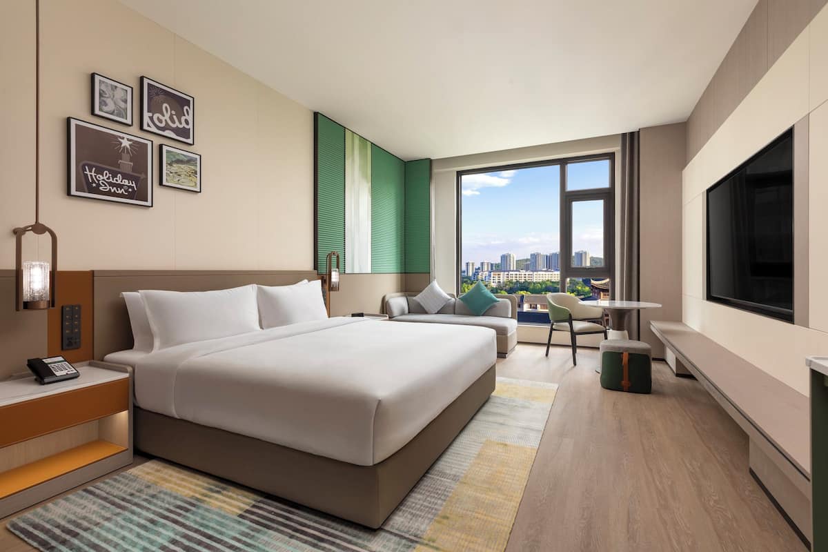 A modern hotel room with a large bed, a desk, a chair, a TV, and a city view.