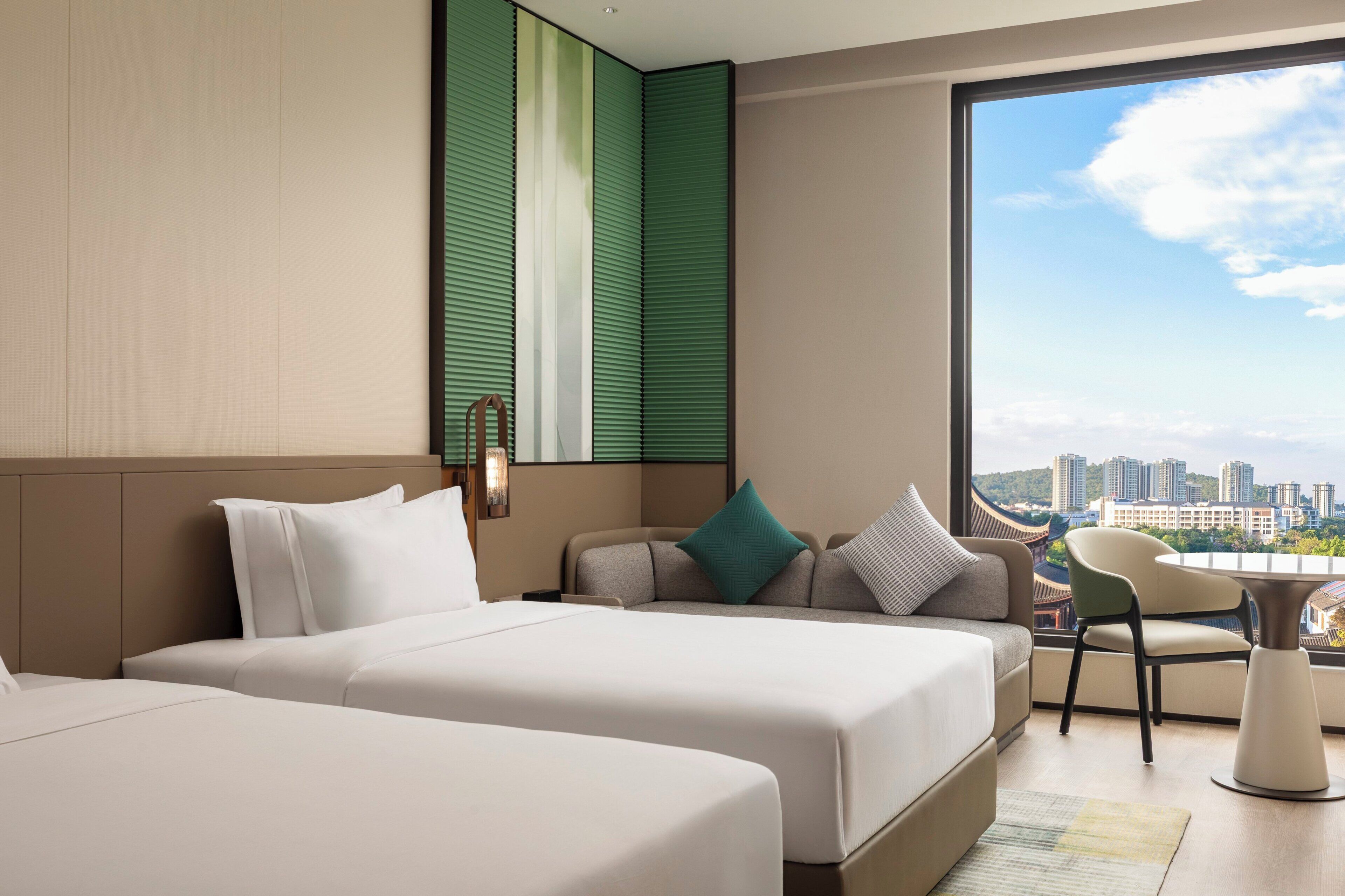 A hotel room with two beds, a sofa, a dining table, and a view of a cityscape.