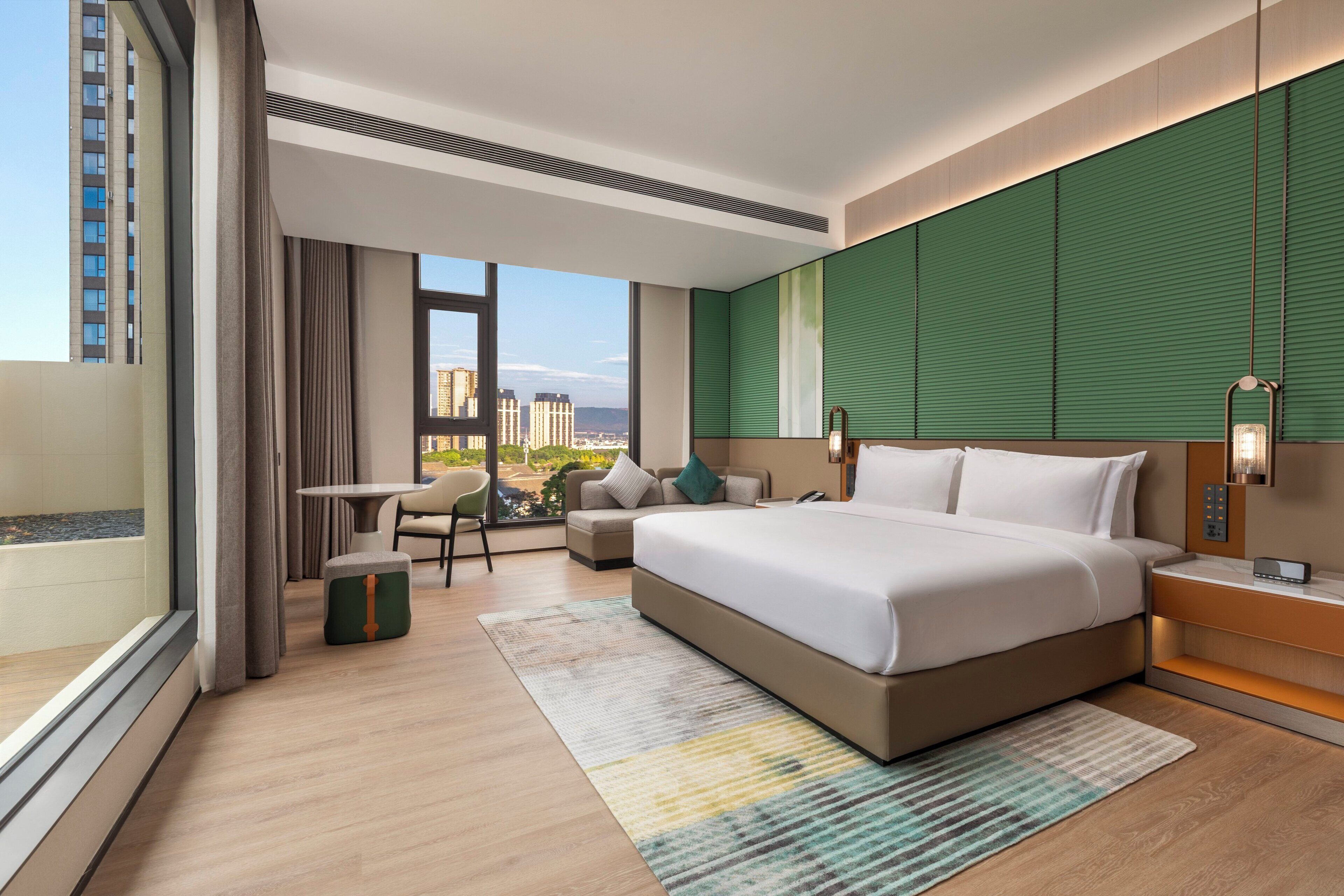 Ein modernes Hotelzimmer mit einem großen Bett, einem kleinen Tisch, einem Sessel und Blick auf die Stadt. Eine grüne Akzentwand setzt ein Farbtupfer.