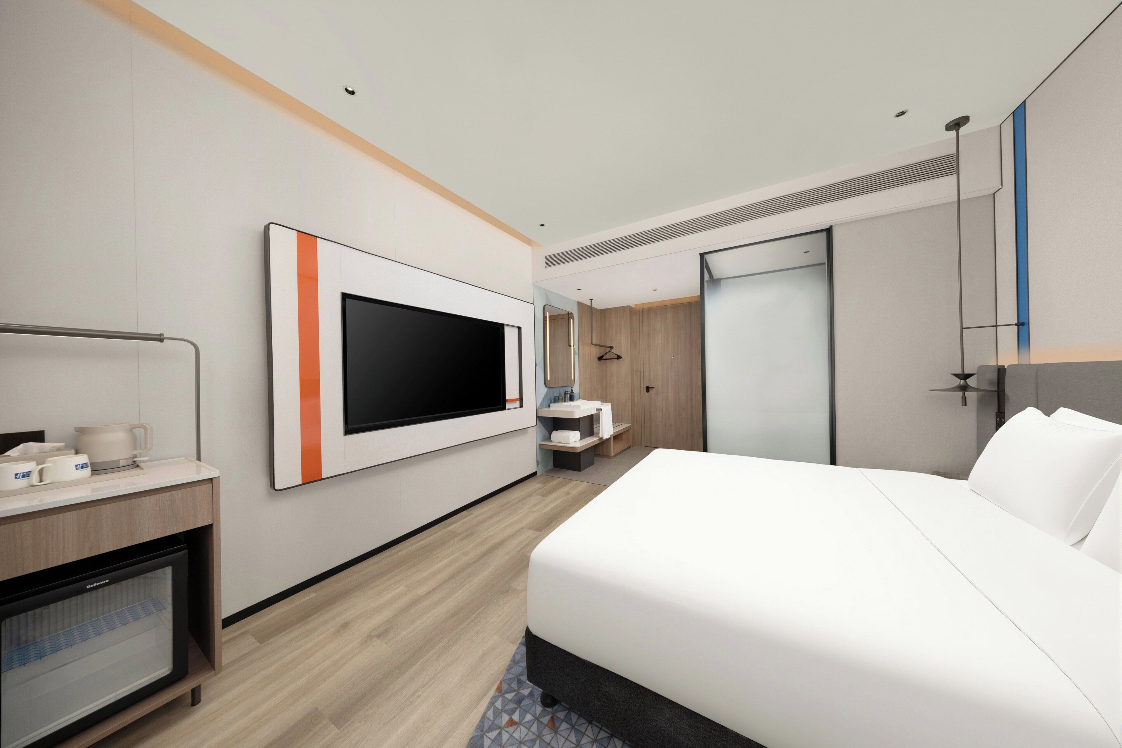 A modern hotel room with a large bed, a flat-screen TV mounted on the wall, a desk with a coffee maker, and a bathroom with a mirror and sink.