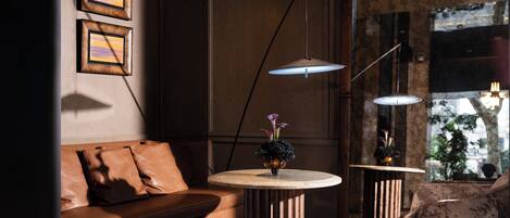 A modern living room with a leather sofa, a round coffee table, and a patterned armchair. There are two pendant lights and a vase with flowers on the table.