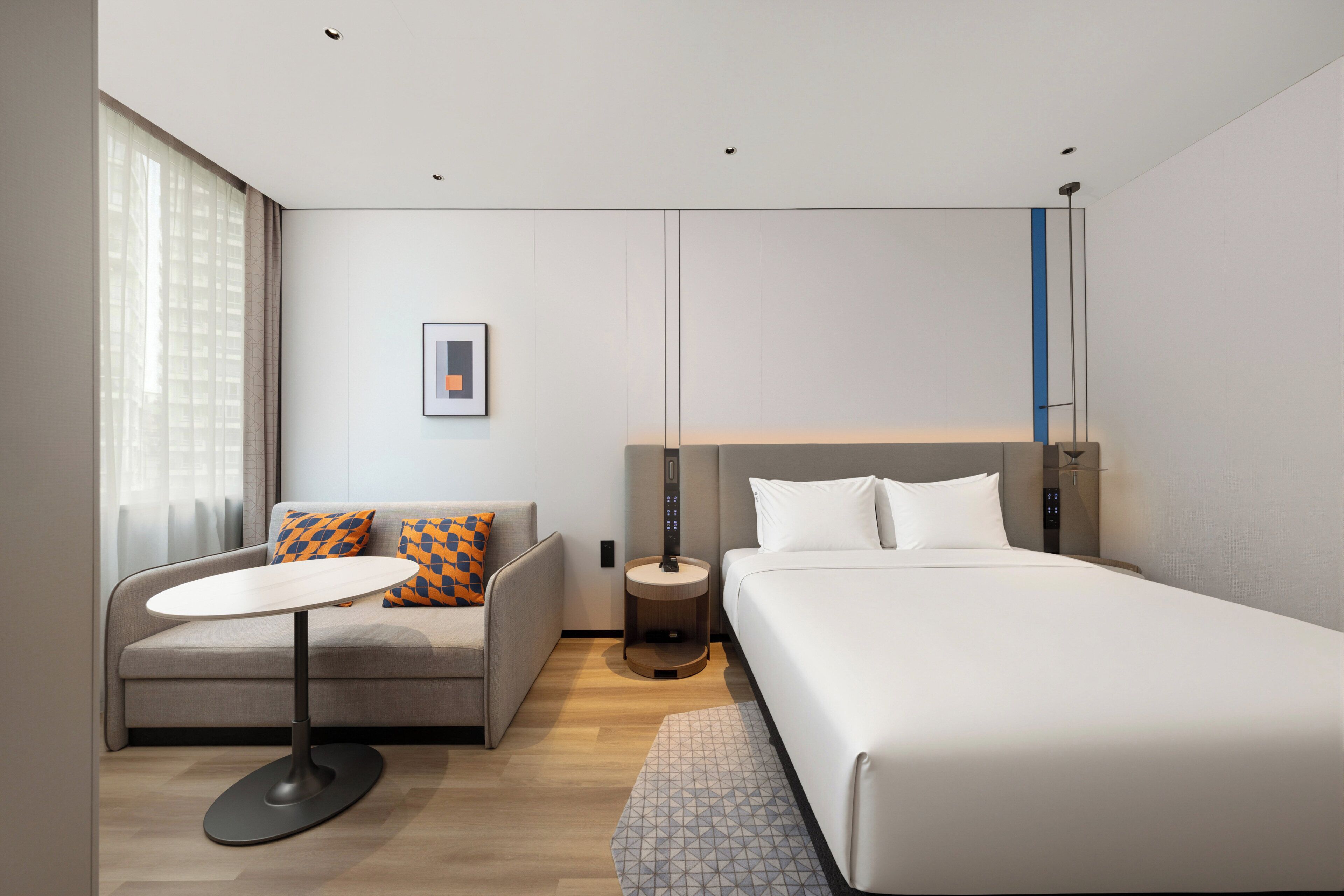 A modern hotel room with a bed, a sofa, a small round table, and a bedside lamp.