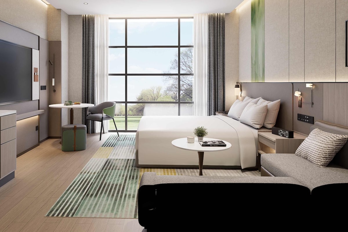 A modern hotel room with a large bed, a desk, a chair, a TV, and a window with a view of trees.