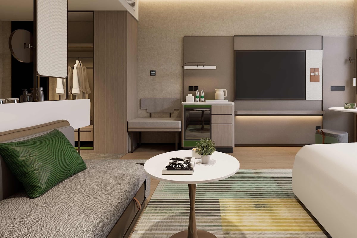 A modern hotel room with a sofa, a small round table, a bed, a kitchenette, and a large window.