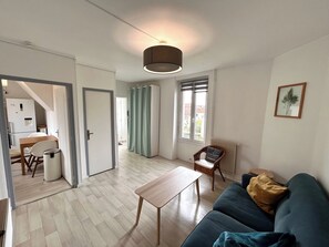 A living room with a sofa, a table, a chair, a radiator, a window with curtains, and a door leading to another room. - Le Millésime Gilbert | Duplex | Calme (Reims)