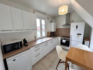 A compact kitchen with white cabinets, a microwave, a sink, a stove, and a dining area with a table and chairs. - Le Millésime Gilbert | Duplex | Calme (Reims)