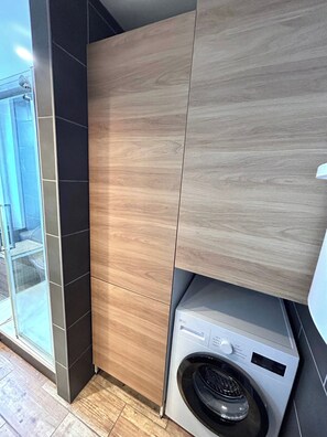 A modern laundry room with a built-in washer and dryer, wooden cabinetry, and a glass door leading to an outdoor area. - La Suite Pierret | Centre-ville (Reims)