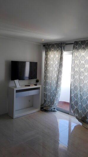 Ein modernes Wohnzimmer mit einem Flachbildfernseher auf einer weißen Konsole, gemusterten Vorhängen und einem großen Fenster.