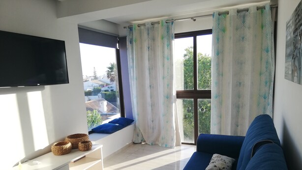 Un salon avec un canapé bleu, un téléviseur fixé au mur, une fenêtre avec des rideaux et une petite table sur laquelle sont posés des paniers.