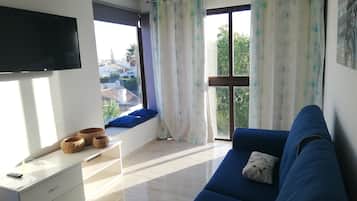 A living room with a blue sofa, a TV mounted on the wall, a window with curtains, and a small table with baskets.