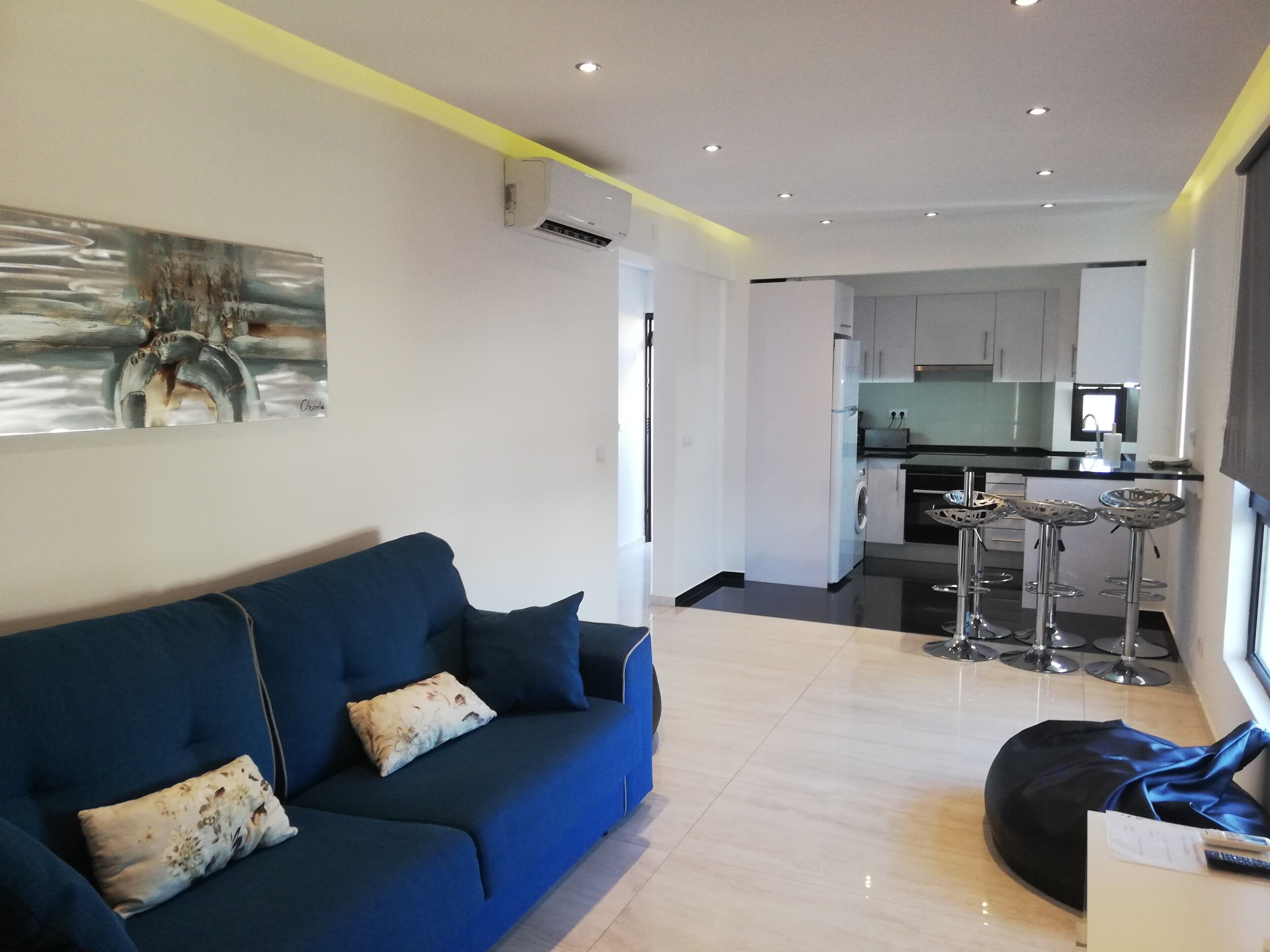 A modern living room with a blue sofa, a black bean bag, and a kitchen in the background.