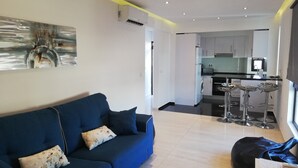 Un salon moderne avec un canapé bleu, un fauteuil en forme de poire noir et une cuisine en arrière-plan.