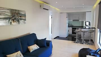 A modern living room with a blue sofa, a black bean bag, and a kitchen in the background.