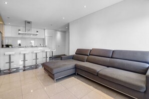 Un espacio de estar moderno con un sofá seccional gris, una cocina con armarios blancos y una zona de bar con taburetes.