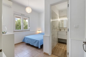 Ein Schlafzimmer mit Bett, Fenster, Nachttischlampe und einem Badezimmer inklusive Spiegel und Waschbecken.