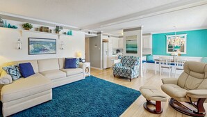 A living room with a sofa, ottoman, and rocking chair, a blue rug, and a kitchen area with a dining table and chairs. - Our Place At The Beach 206G (Ocean City)