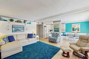 Ein Wohnzimmer mit Sofa, Hocker und Schaukelstuhl, einem blauen Teppich und einer Küchenzeile mit Esstisch und Stühlen.
