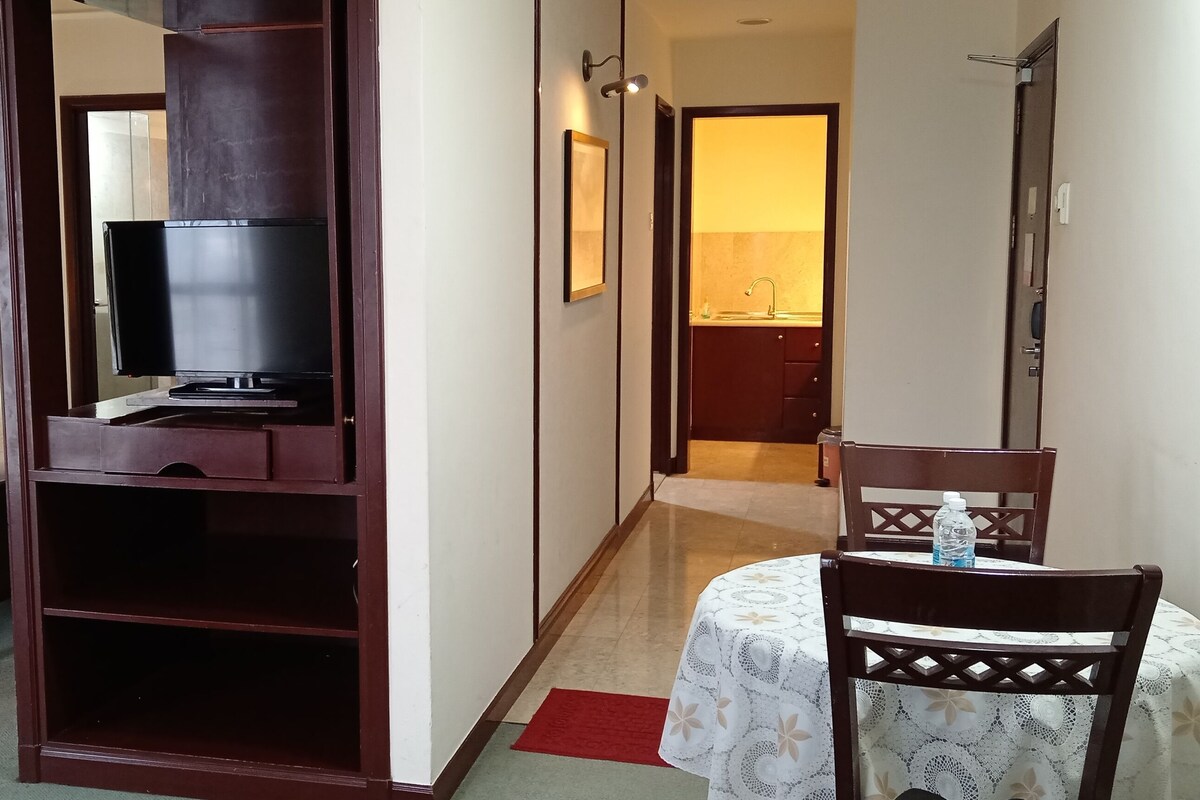 Quarto de hotel com cama, televisão, escrivaninha com cadeira, espelho e banheiro visível através de uma porta aberta.