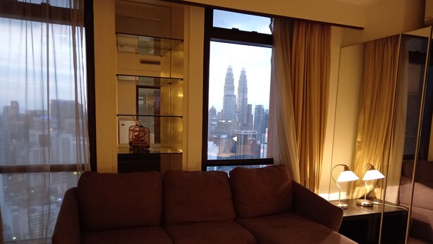 A modern living room with a brown sofa, a glass cabinet, and a view of the cityscape including the Petronas Towers. - Berjaya Times Square Apartment (Kuala Lumpur)
