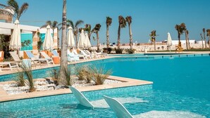A poolside area with lounge chairs and umbrellas, palm trees, and a clear blue sky. - Makai Hotel North Coast By Brassbell (El Dabaa)