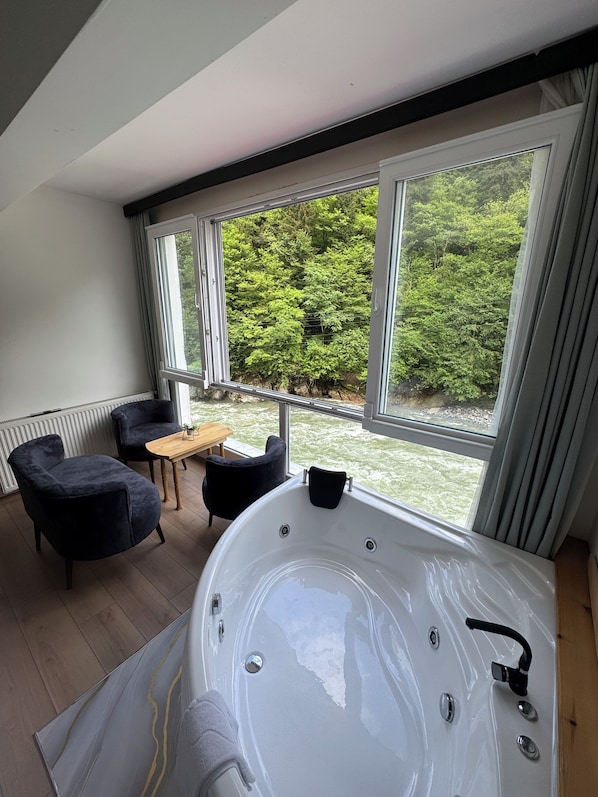 A modern bathroom with a large bathtub, two dark armchairs, a wooden table, and a view of a river and forest through large windows.