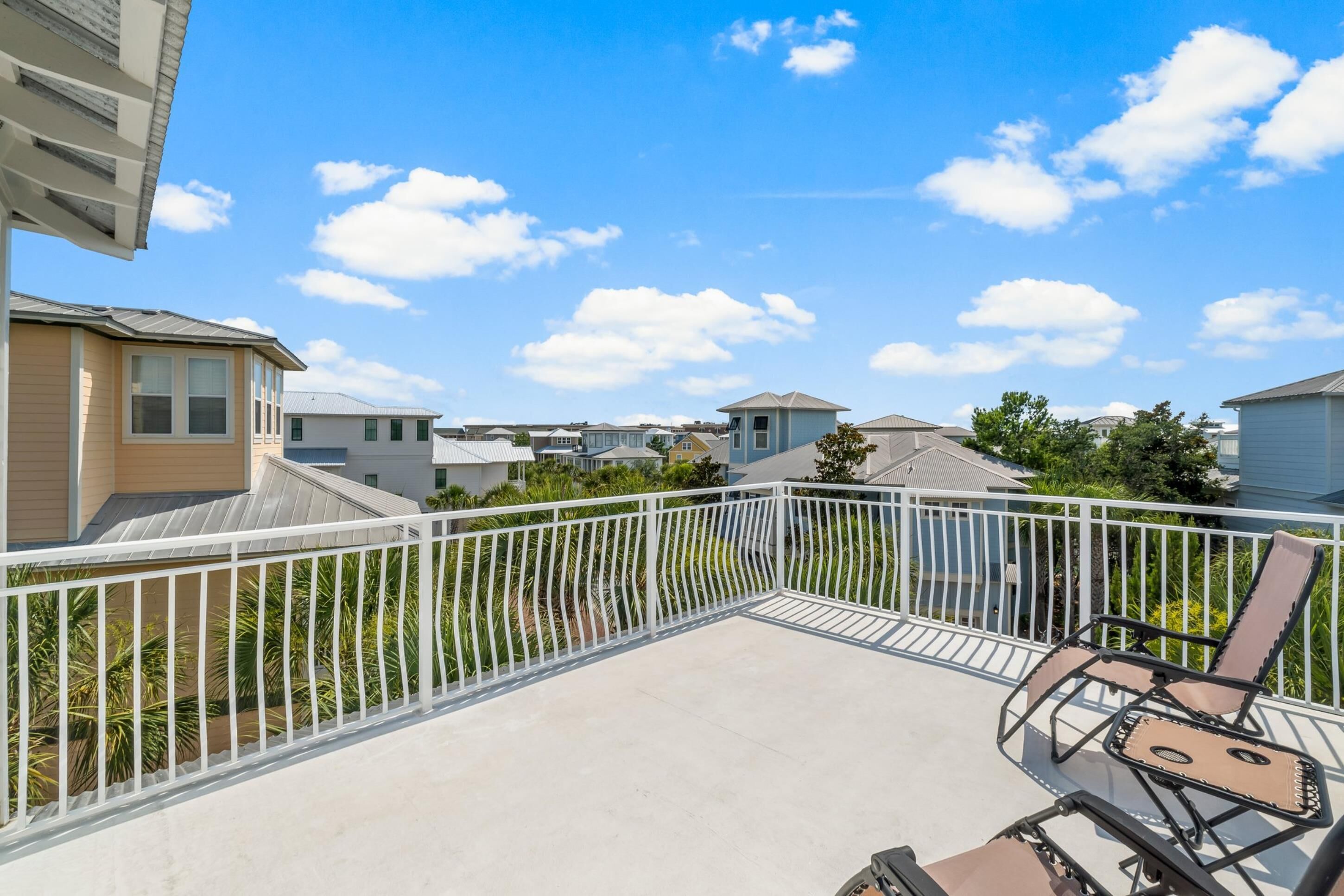 Un balcon d’habitation avec une balustrade blanche, deux fauteuils confortables et une vue sur les maisons voisines, le tout sous un ciel dégagé.
