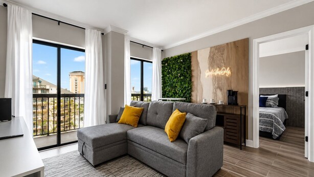 Un appartement moderne avec un canapé d’angle gris, un petit bureau, une machine à café, un lit avec une tête de lit grise et un mur vert agrémenté d’un jardin vertical.