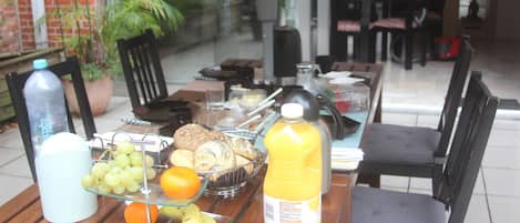 A dining table with a glass fruit bowl containing grapes, an orange, and bananas, a bottle of orange juice, and a water bottle.
