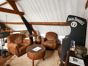 Una habitación estilo loft con vigas de madera a la vista, dos sillones de cuero, una pequeña mesa de madera y un barril. Se observa un letrero negro con 'Jack Daniel's'.
