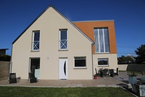 A modern two-story house with a flat roof, a front door, and a small patio area with outdoor furniture.