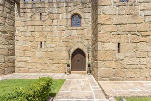 A stone building with a large arched entrance, a small window with a decorative grille, and a paved pathway leading to the entrance.