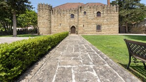 Um edifício de pedra com entrada central, um caminho pavimentado que leva a ele e um banco à direita.