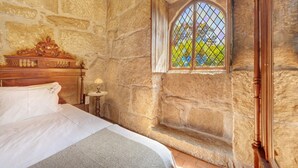 Quarto com paredes de pedra, uma cama, cabeceira de madeira, uma mesa pequena com abajur e uma janela com vista para o verde.