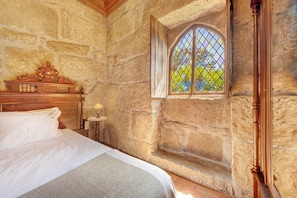 Une pièce aux murs de pierre, avec un lit, une tête de lit en bois, une petite table avec une lampe et une fenêtre donnant sur un paysage verdoyant.