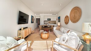 A modern living room with a flat-screen TV, a sofa, a dining area, and a kitchen in the background. - Casa da Saudade in Tavira (Tavira)
