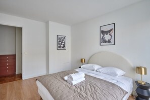 Ein ordentlich bezogenes Bett mit einer beigen Steudecke, weißen Kissen und einem gefalteten Handtuch. Zwei Nachttische mit Lampen stehen bereit, und der Raum hat eine weiße Tür und Wände.