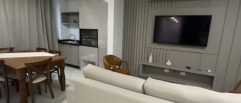 Una sala de estar moderna con sofá, mesa de comedor, sillas, zona de cocina con un lavamanos y un televisor de pared.