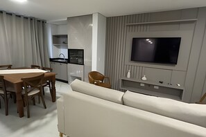 A modern living room with a sofa, dining table, chairs, a kitchen area with a sink, and a mounted TV. - Apartamento no centro de Foz do Iguaçu (Foz do Iguaçu)